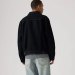 A man viewed from behind wearing a black denim jacket over a black and white striped shirt, paired with light-wash blue jeans, against a plain light gray studio background.