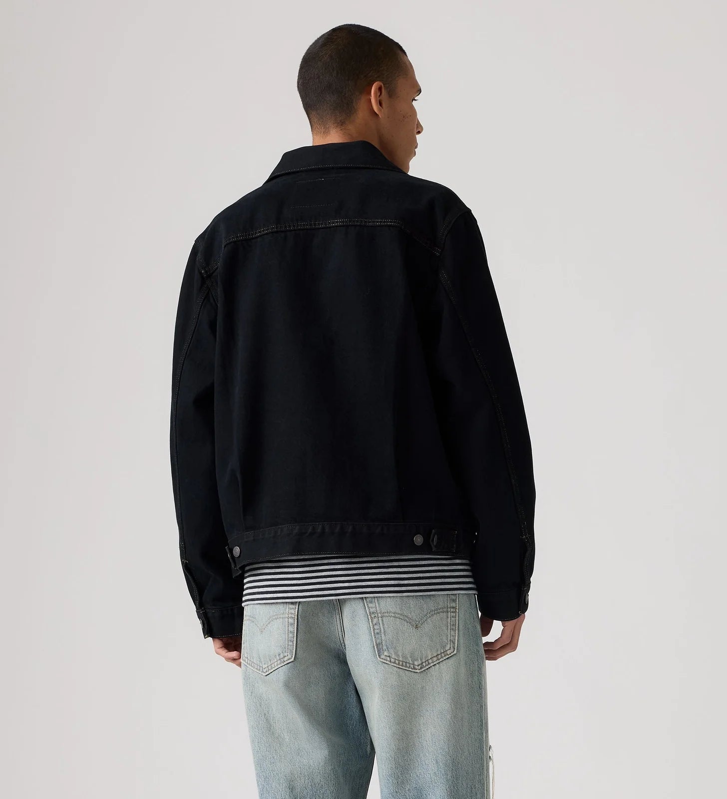 A man viewed from behind wearing a black denim jacket over a black and white striped shirt, paired with light-wash blue jeans, against a plain light gray studio background.