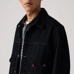 A close-up of a model wearing a dark navy denim trucker jacket over a black and white striped t-shirt, against a light gray studio background.