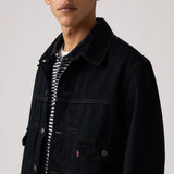A close-up of a model wearing a dark navy denim trucker jacket over a black and white striped t-shirt, against a light gray studio background.