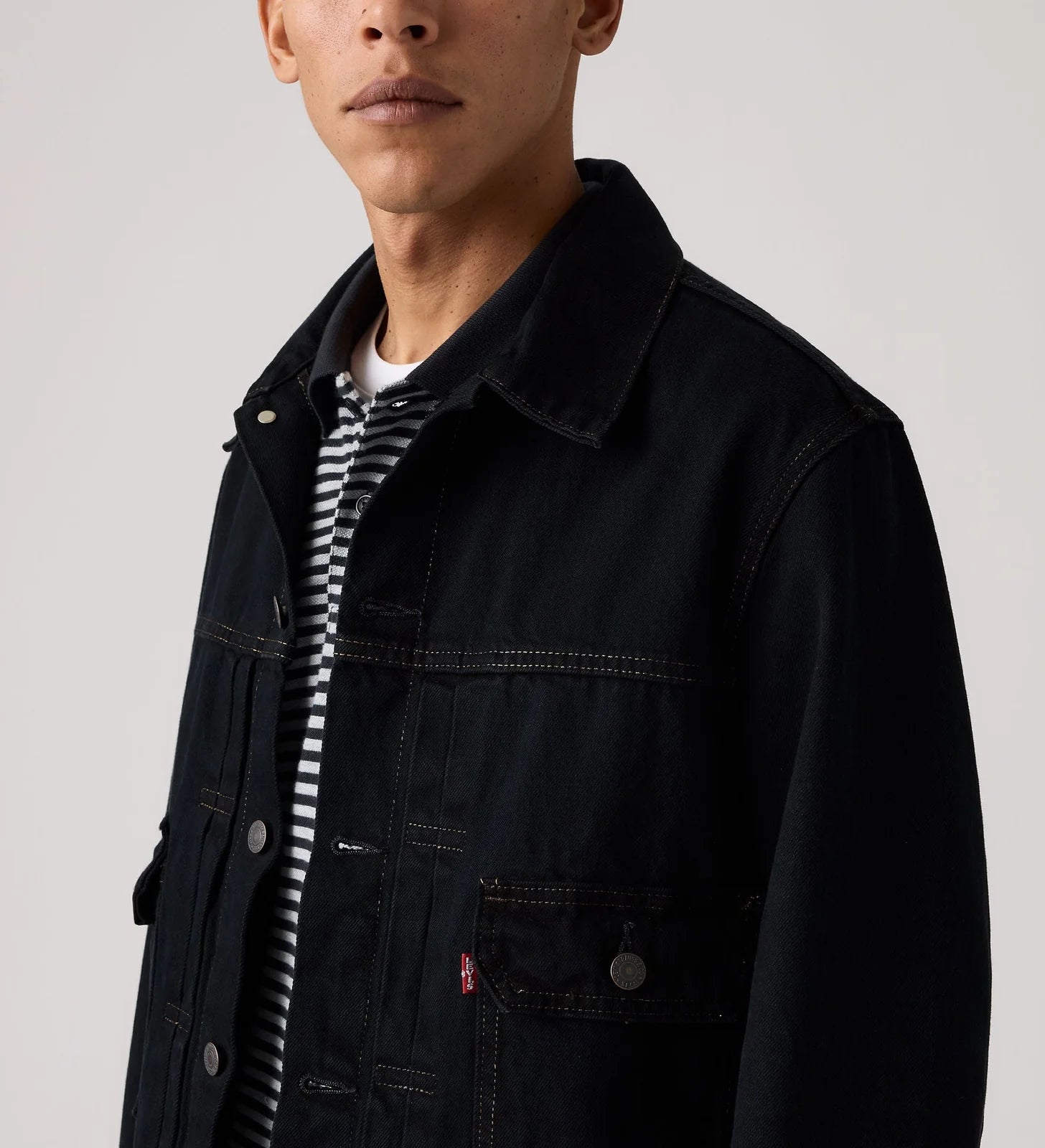 A close-up of a model wearing a dark navy denim trucker jacket over a black and white striped t-shirt, against a light gray studio background.