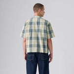 A man viewed from the back wearing a short-sleeve plaid button-up shirt in cream, green, and blue, paired with dark blue jeans, against a light gray studio background.