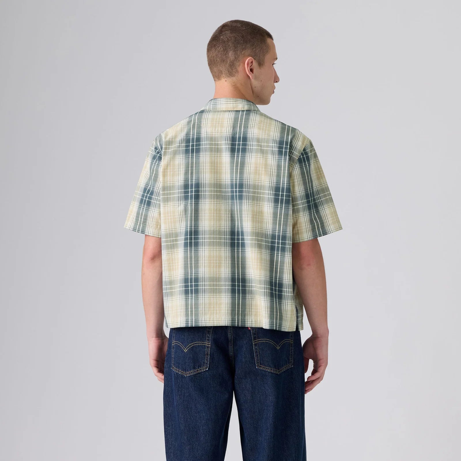 A man viewed from the back wearing a short-sleeve plaid button-up shirt in cream, green, and blue, paired with dark blue jeans, against a light gray studio background.