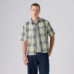 A young man modeling a short-sleeve button-up plaid shirt in green, beige, and white, worn open over a beige t-shirt and dark blue jeans, against a light gray studio background.