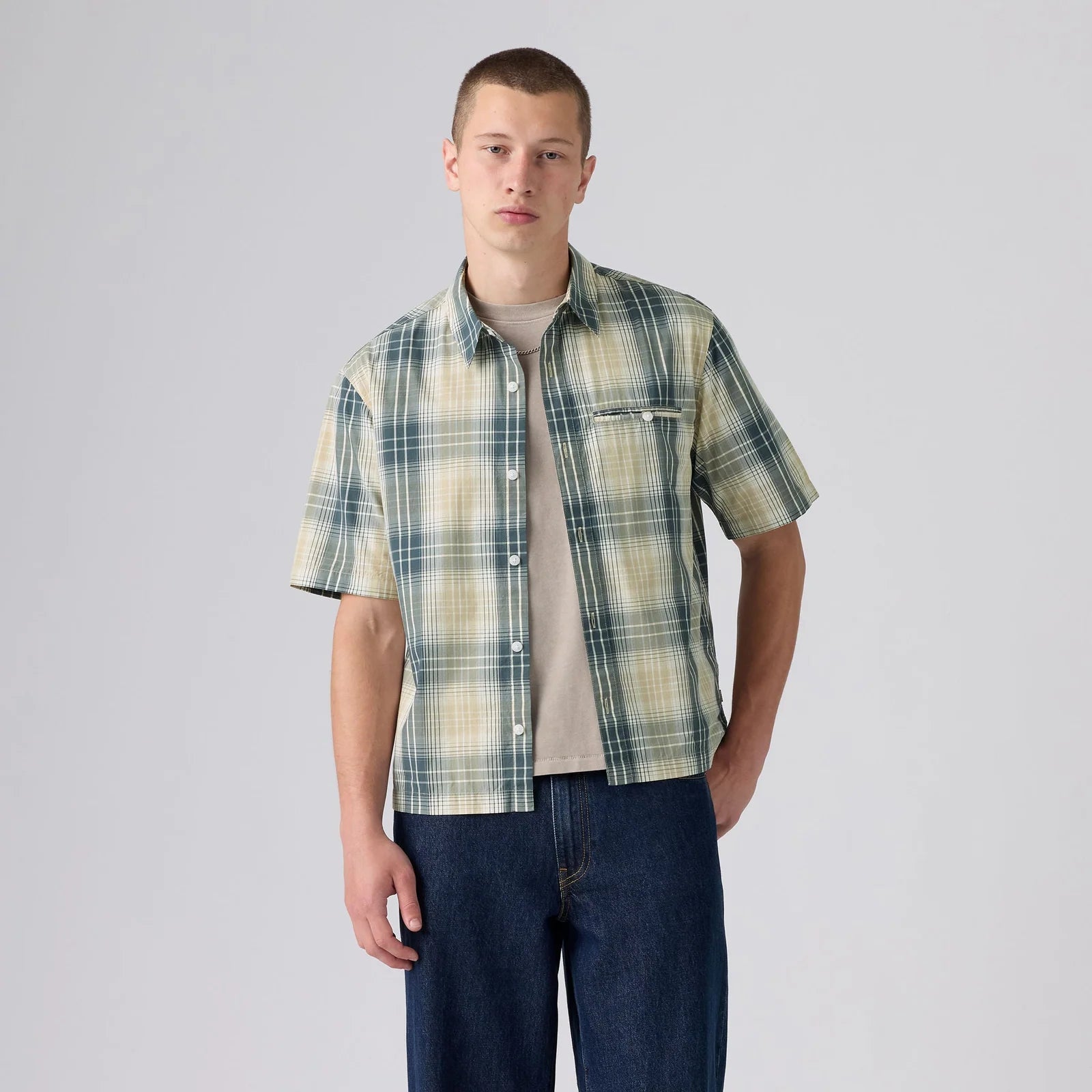 A young man modeling a short-sleeve button-up plaid shirt in green, beige, and white, worn open over a beige t-shirt and dark blue jeans, against a light gray studio background.