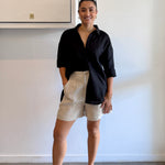 Woman wearing a black shirt and beige shorts standing against a white wall.