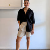 Woman wearing a black shirt and beige shorts standing against a white wall.