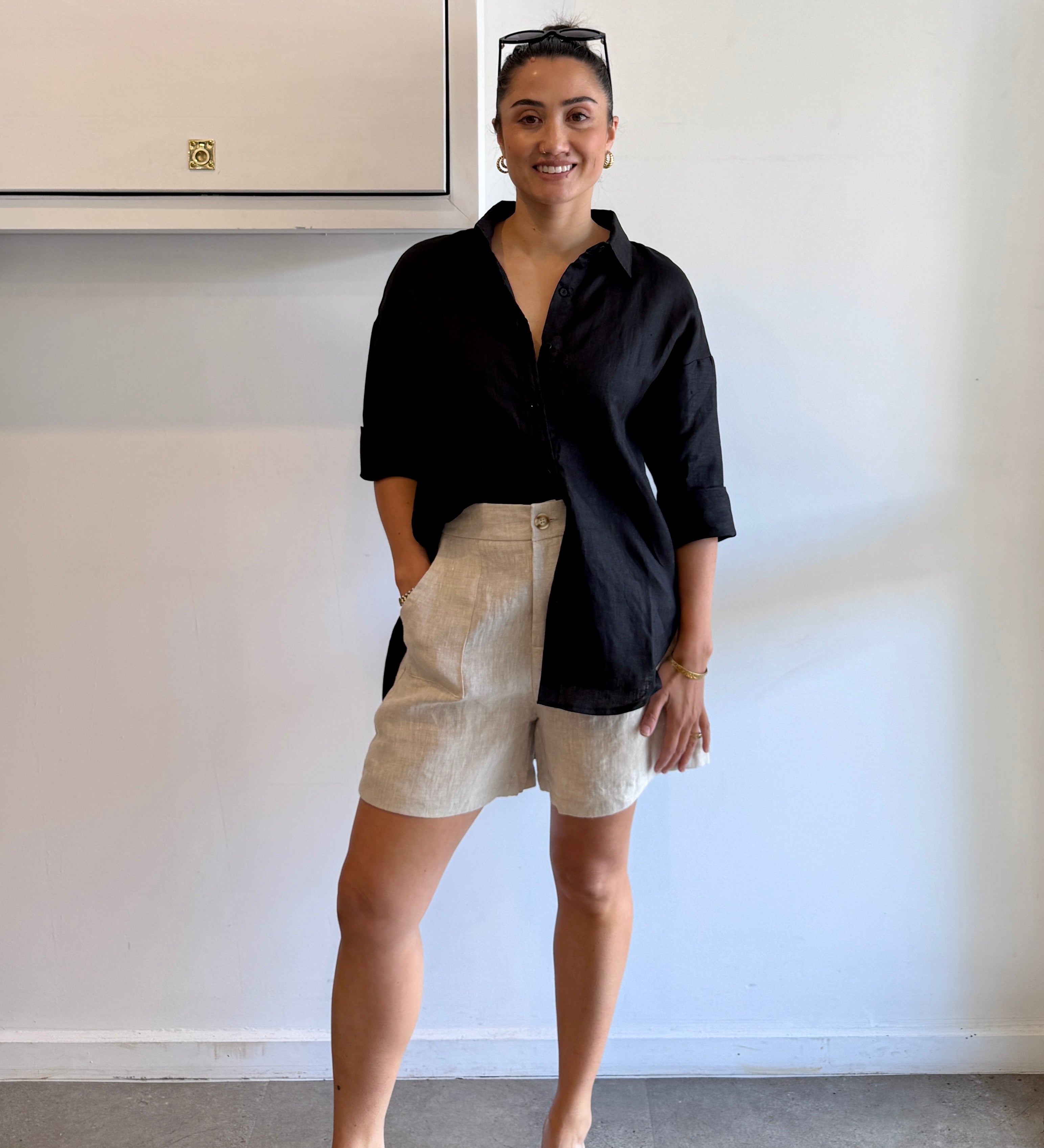 Woman wearing a black shirt and beige shorts standing against a white wall.