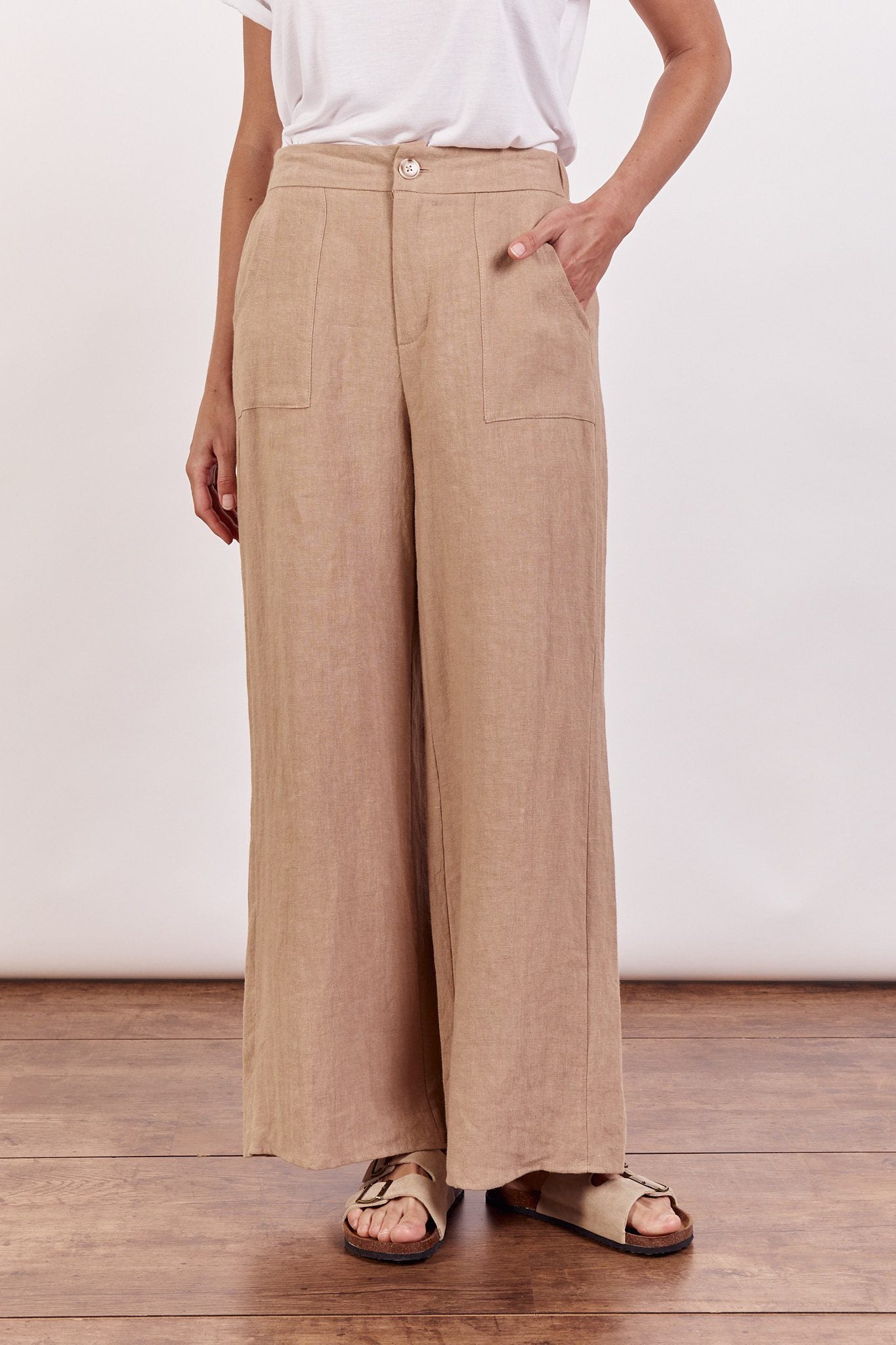 A woman standing in a taupe linen wide-leg pant with a white top, wearing flat sandals.