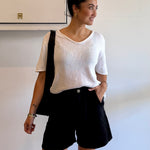 Woman wearing a white top and black shorts with a black bag, standing against a plain wall.