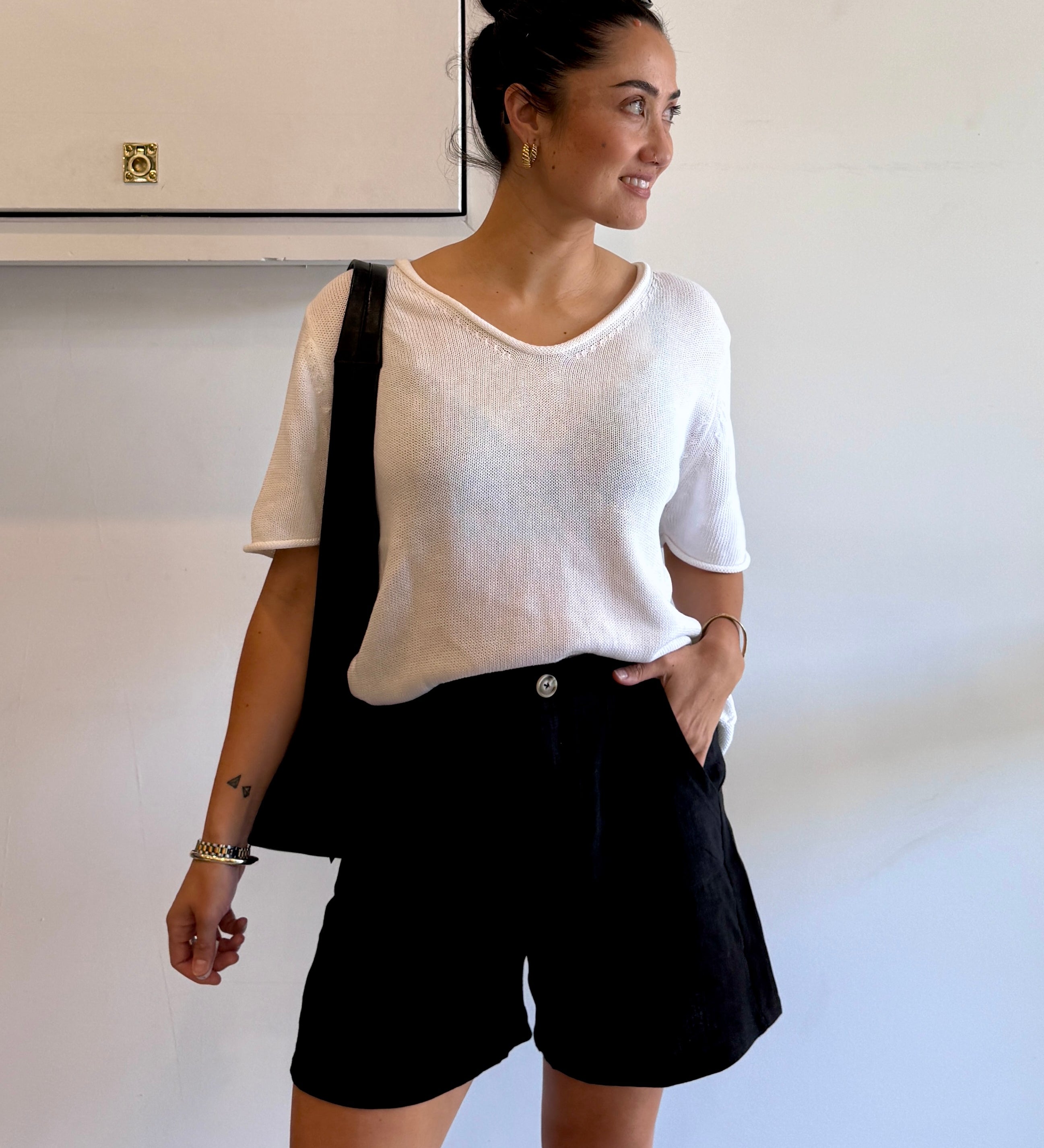 Woman wearing a white top and black shorts with a black bag, standing against a plain wall.
