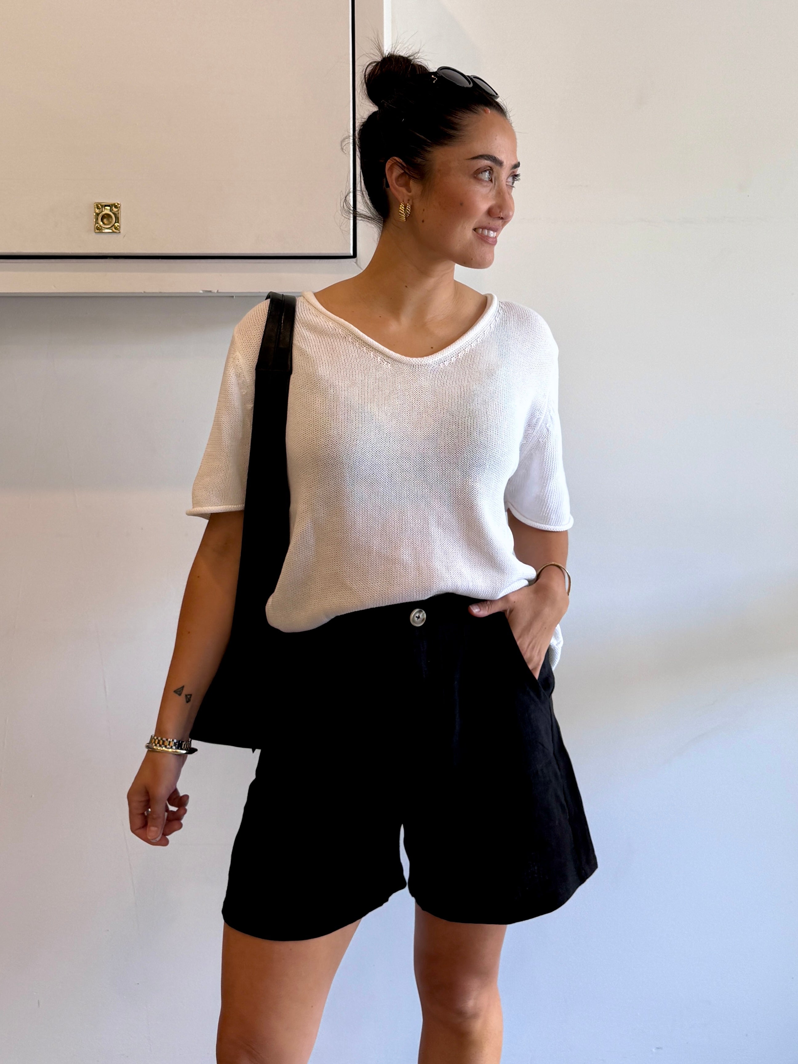 Woman wearing a white top and black shorts with a black bag, standing against a plain wall.