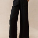 A person standing in a neutral pose wearing black wide-leg linen pants and black sandals, against a beige background.