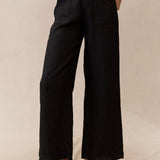 A person standing in a neutral pose wearing black wide-leg linen pants and black sandals, against a beige background.