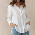 A woman wearing a white, collared, button-up shirt with cuffed sleeves and a high/low hemline, paired with blue denim pants.
