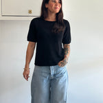 Woman wearing a black t-shirt and light blue jeans standing against a white wall.