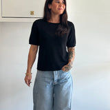 Woman wearing a black t-shirt and light blue jeans standing against a white wall.