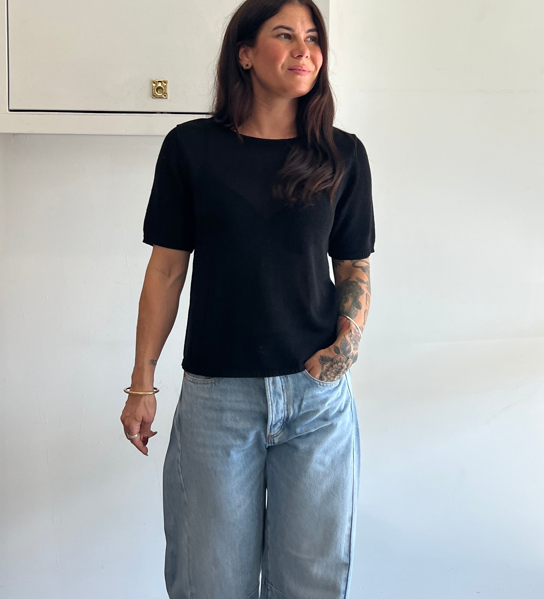 Woman wearing a black t-shirt and light blue jeans standing against a white wall.