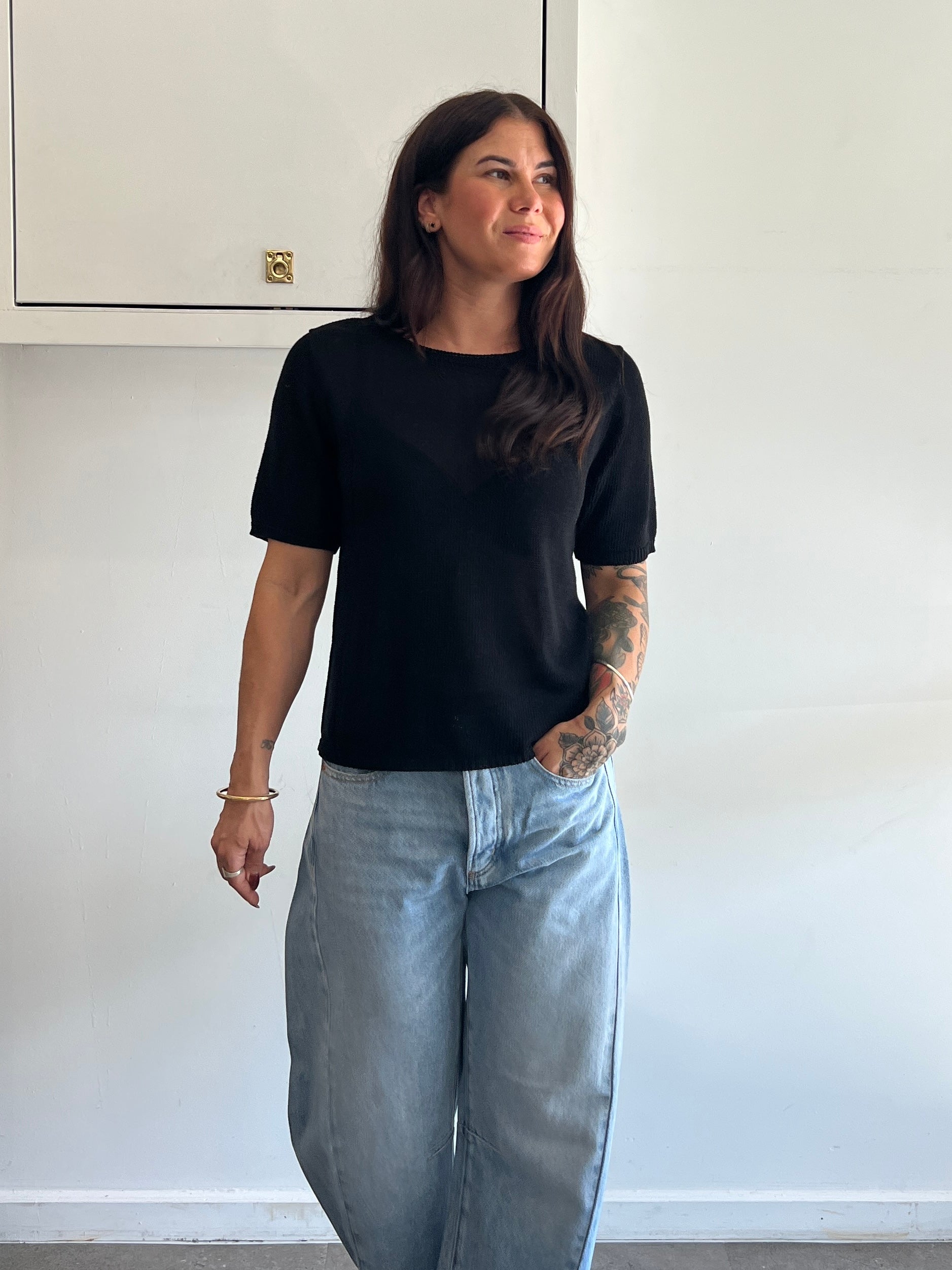 Woman wearing a black t-shirt and light blue jeans standing against a white wall.