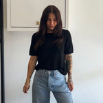 Person wearing a black t-shirt and light blue jeans standing against a white wall.