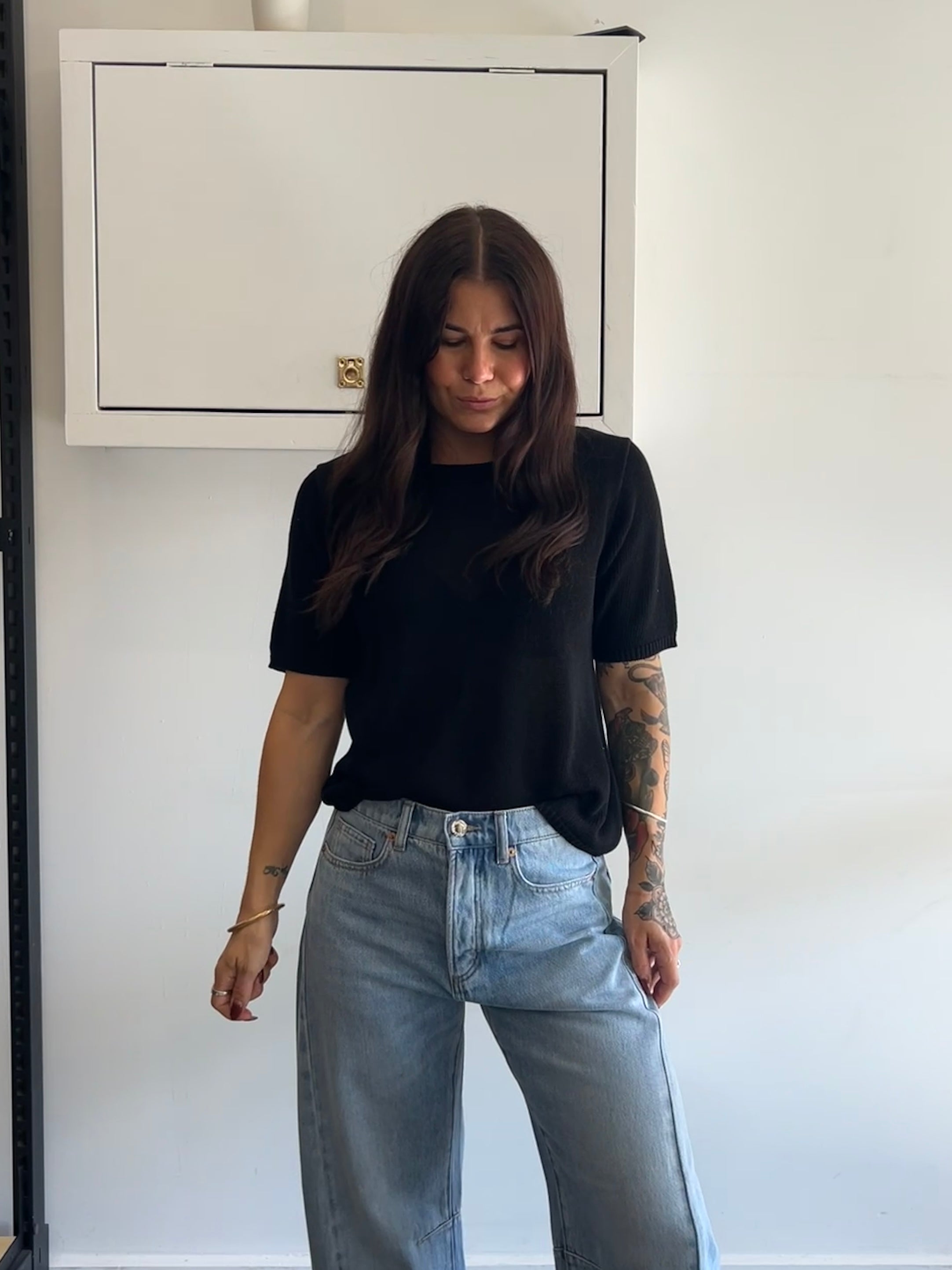 Person wearing a black t-shirt and light blue jeans standing against a white wall.