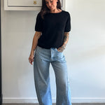 Person wearing a black t-shirt and light blue jeans standing against a white wall.
