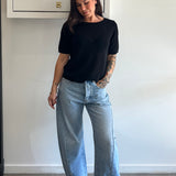 Person wearing a black t-shirt and light blue jeans standing against a white wall.