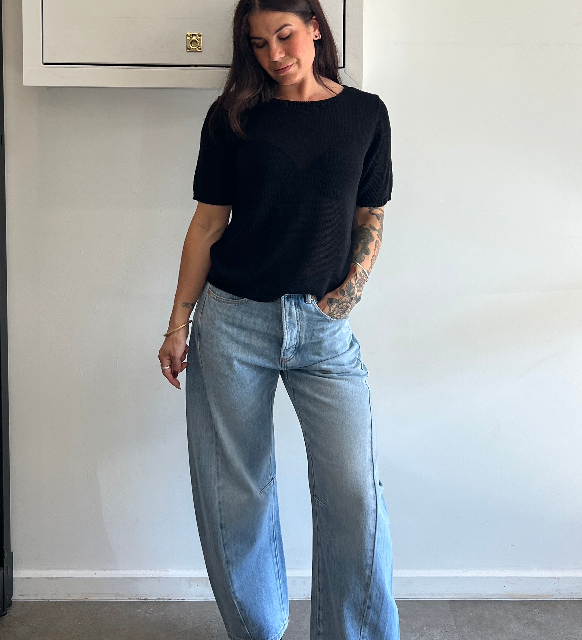 Person wearing a black t-shirt and light blue jeans standing against a white wall.