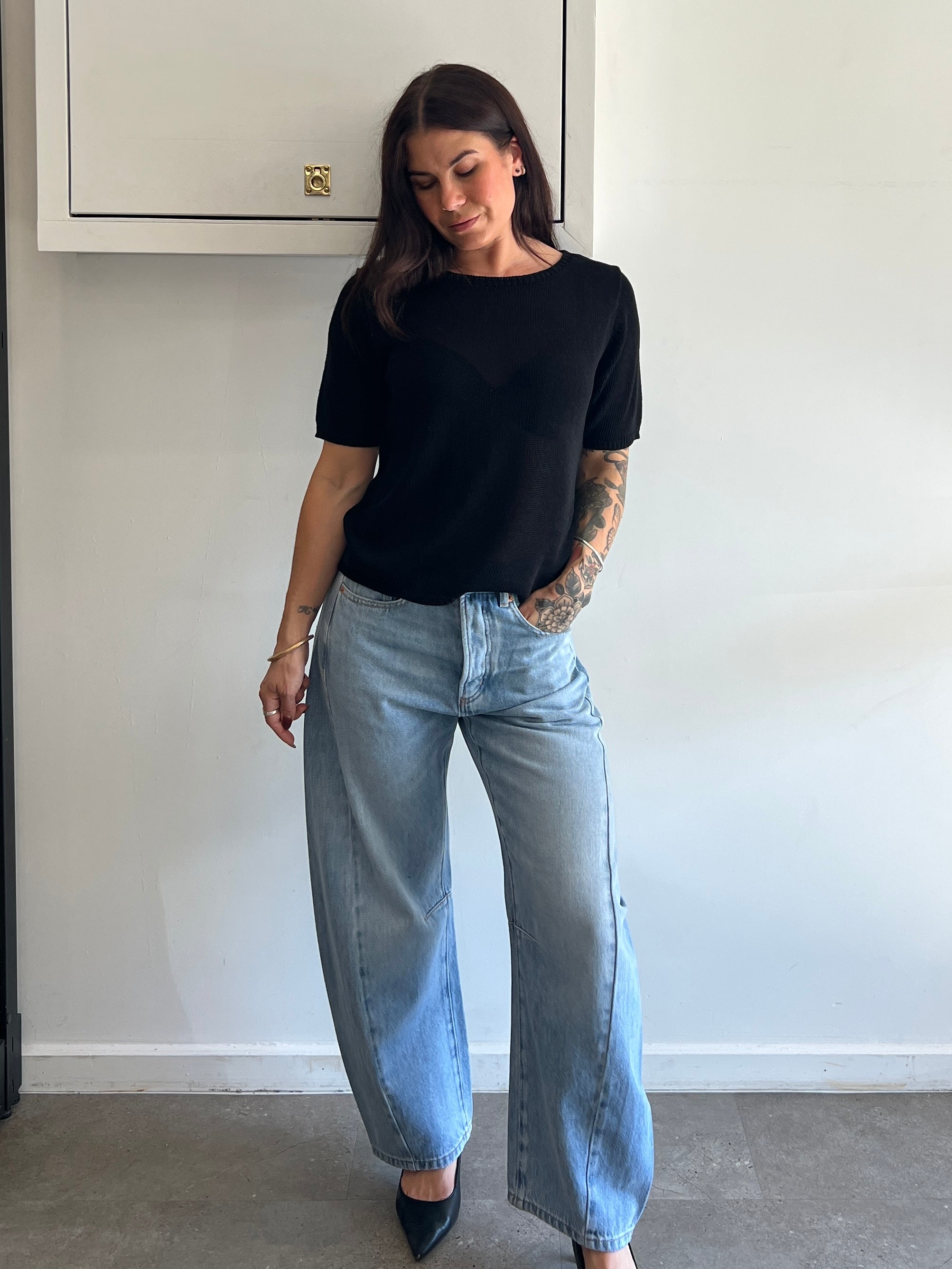Person wearing a black t-shirt and light blue jeans standing against a white wall.
