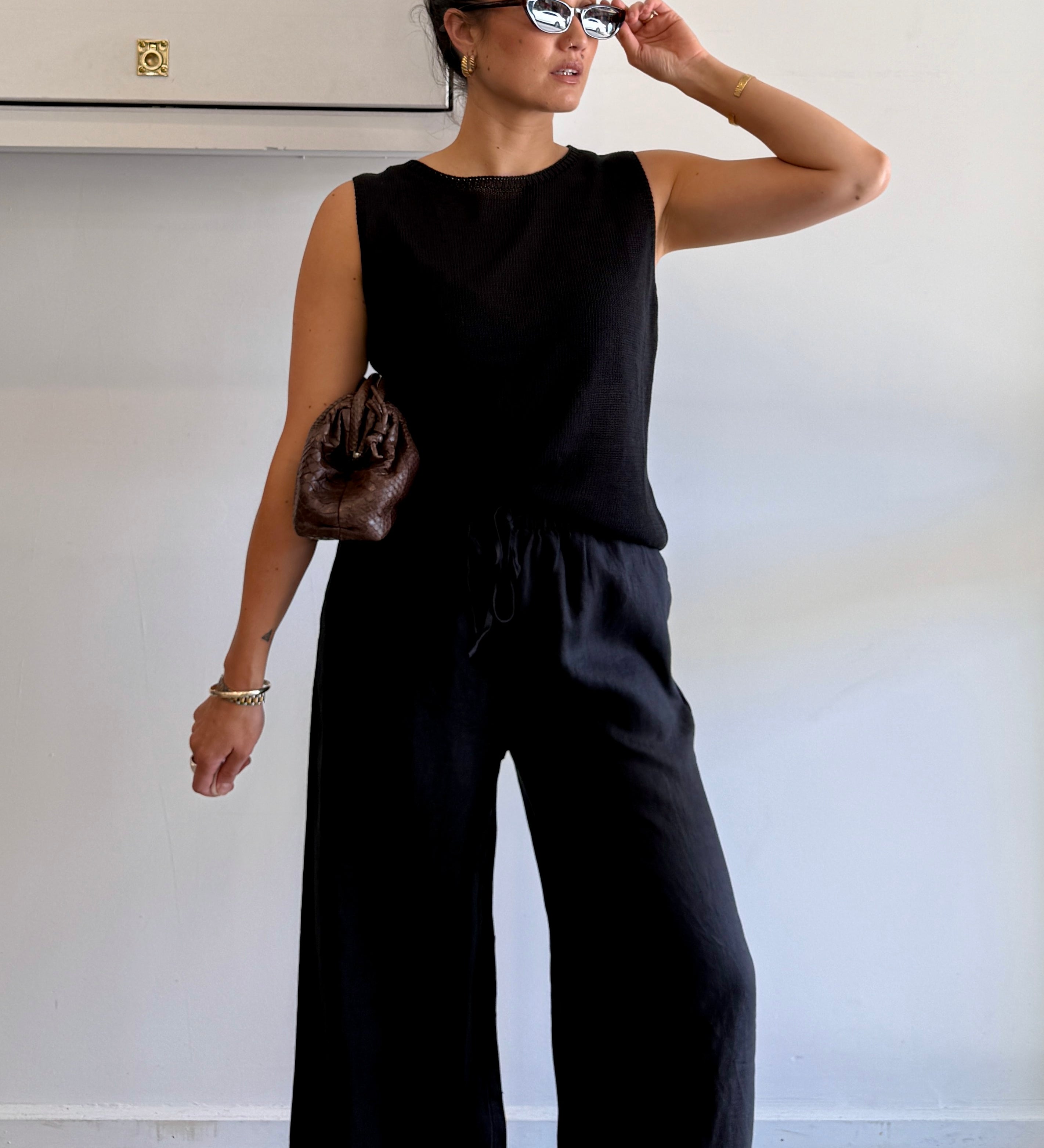 Woman wearing a black sleeveless top and wide-leg pants, holding sunglasses.