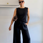 Woman wearing a black sleeveless top and wide-leg pants against a plain background