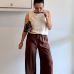 Woman wearing a beige sleeveless top and brown pants, standing against a plain wall.