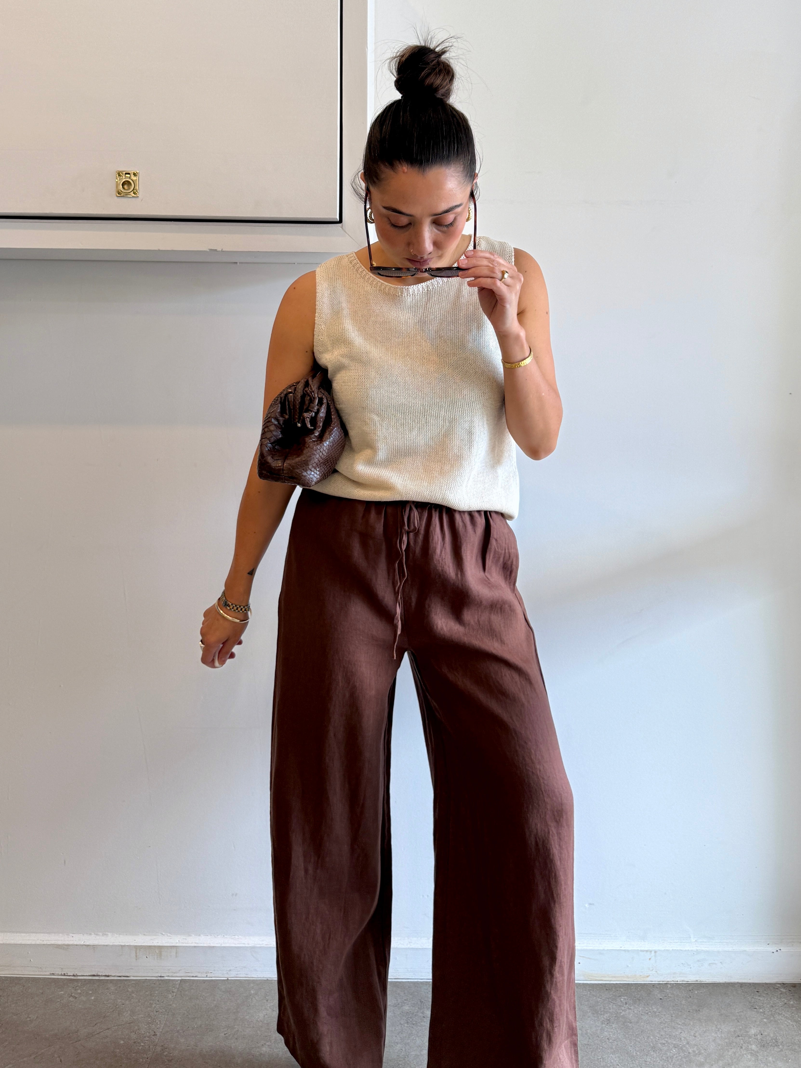 Woman wearing a beige sleeveless top and brown pants, standing against a plain wall.