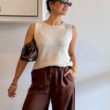 Woman wearing a beige sleeveless top and brown pants against a white wall.