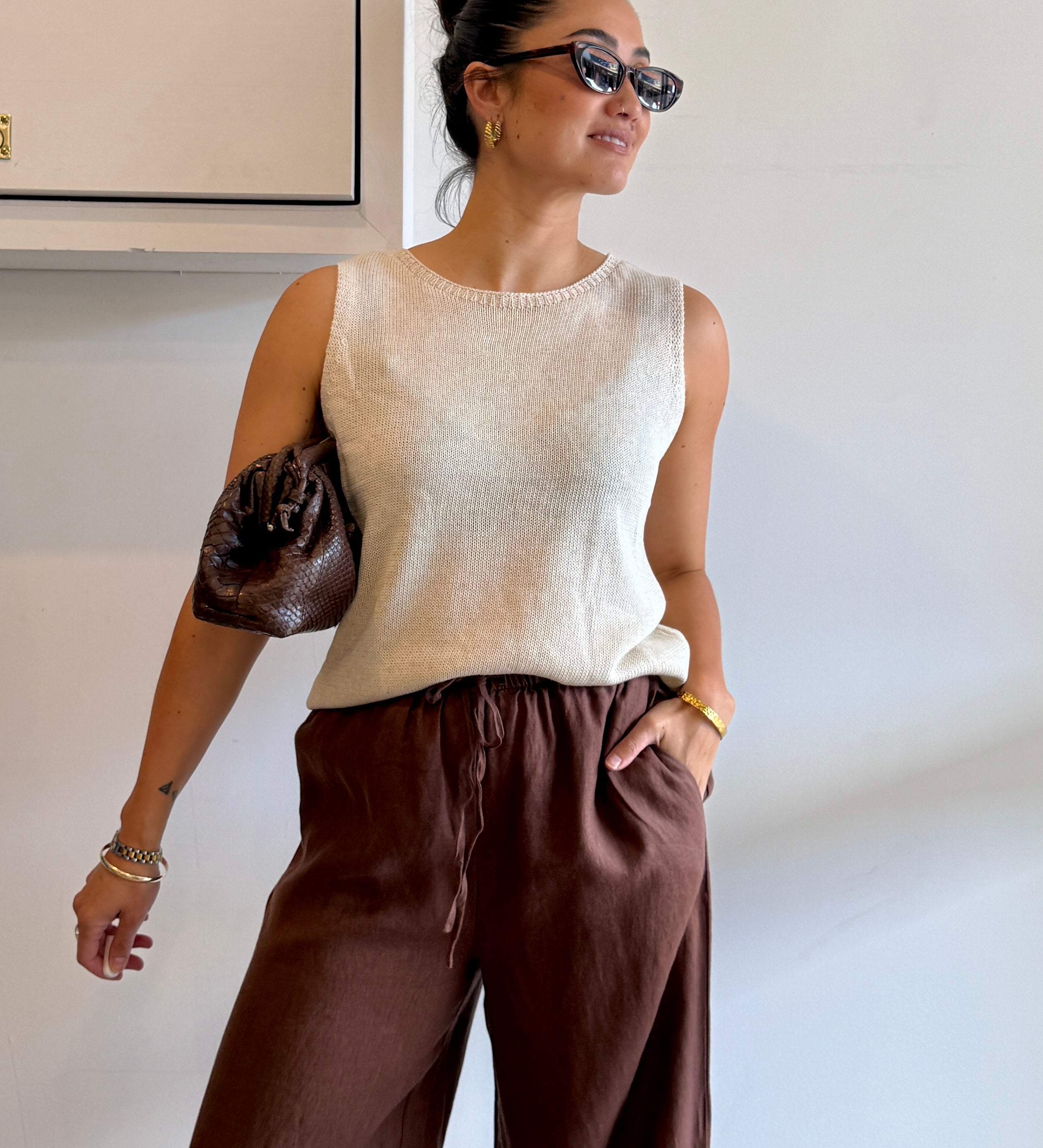 Woman wearing a beige sleeveless top and brown pants against a white wall.
