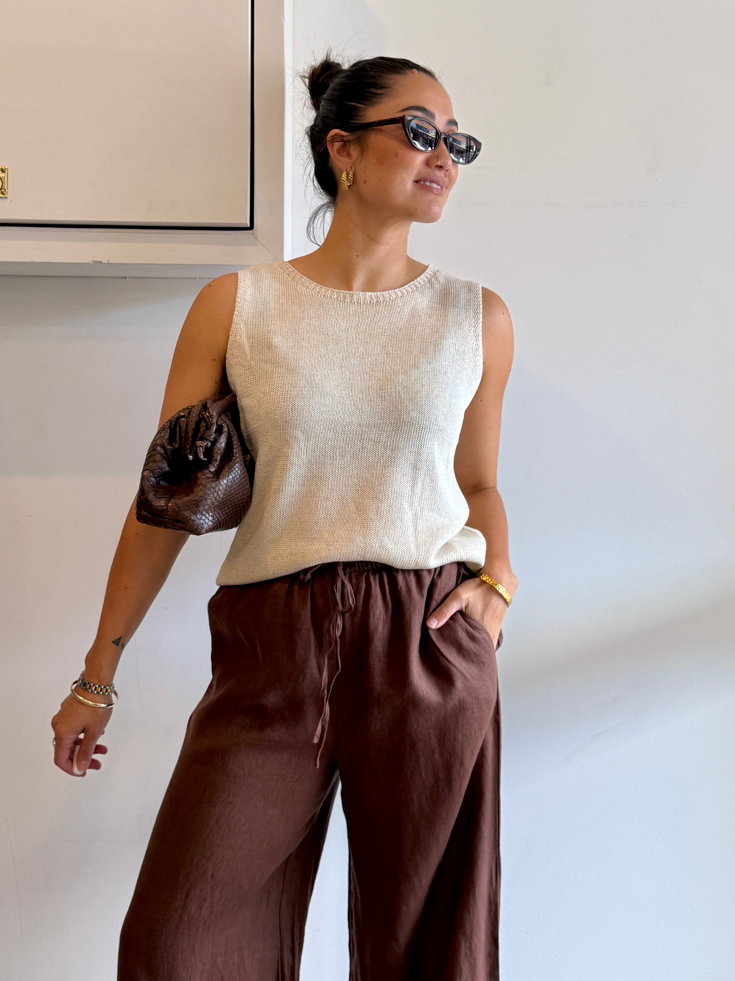 Woman wearing a beige sleeveless top and brown pants against a white wall.