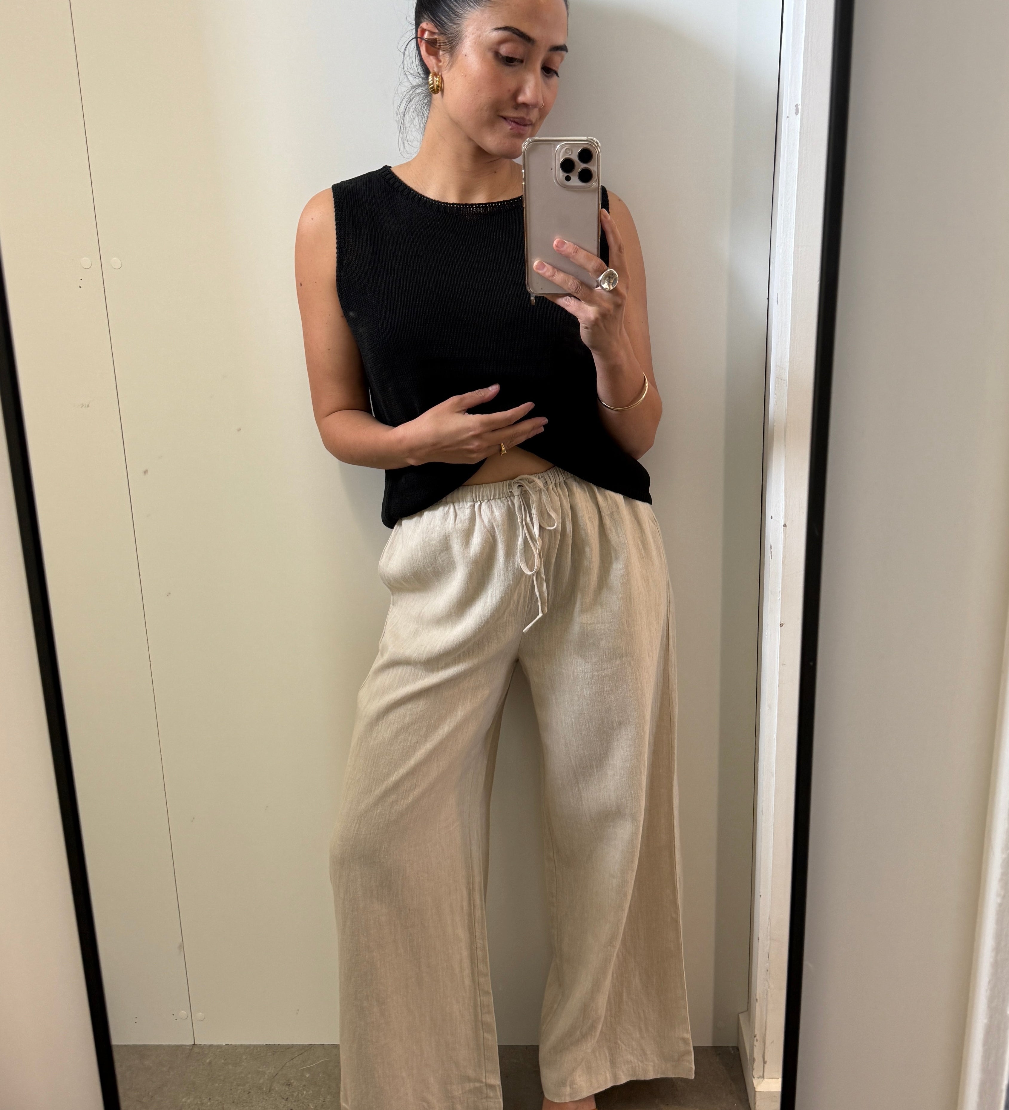 Woman taking a mirror selfie wearing a black sleeveless top and beige pants.