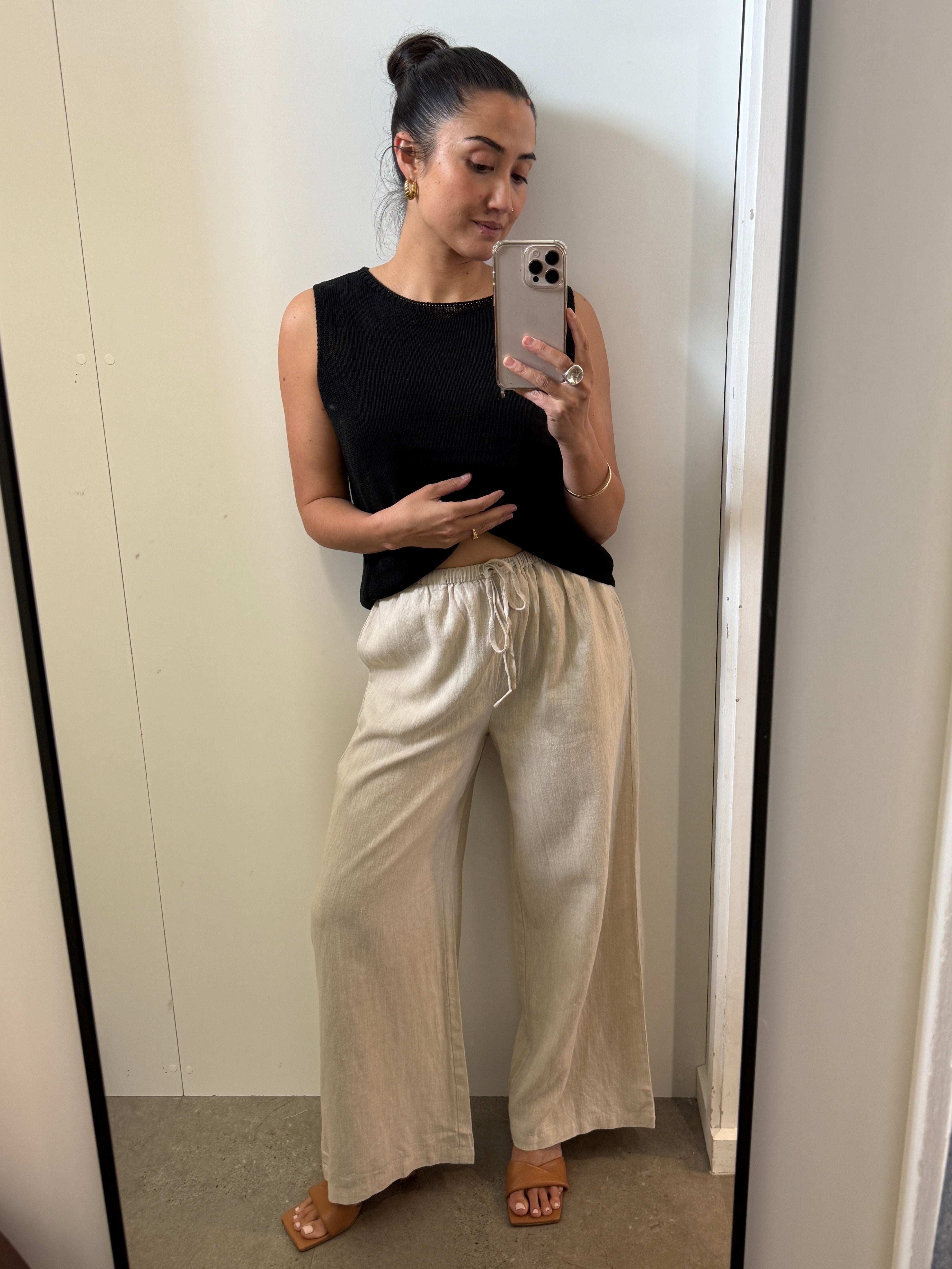 Woman taking a mirror selfie wearing a black sleeveless top and beige pants.