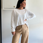 Woman wearing a white long sleeve top and beige pants against a plain wall.