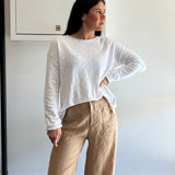Woman wearing a white long sleeve top and beige pants against a plain wall.