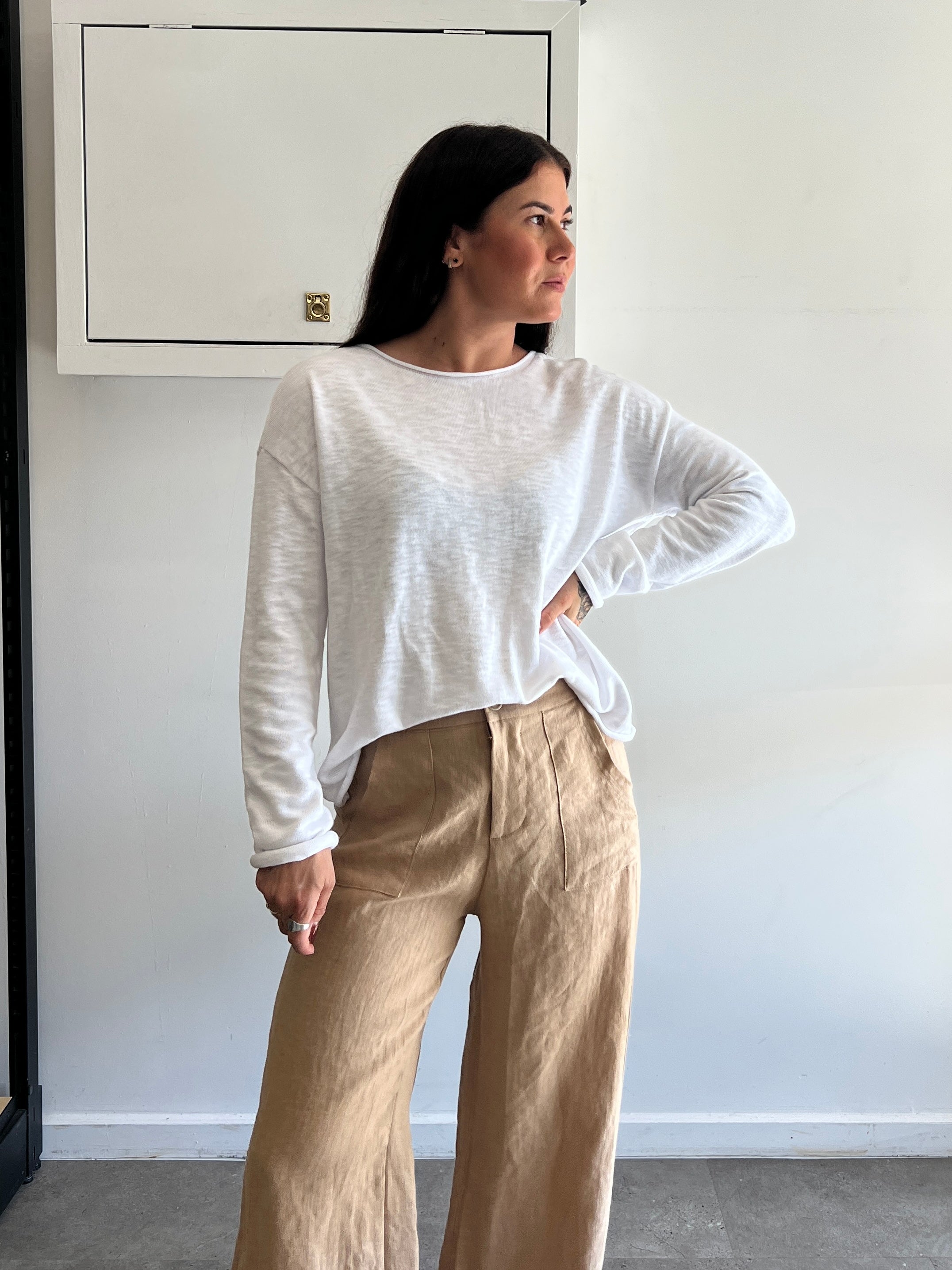 Woman wearing a white long sleeve top and beige pants against a plain wall.