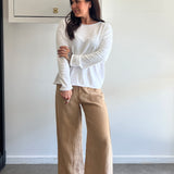 Woman wearing a white long-sleeve top and beige pants against a white wall.