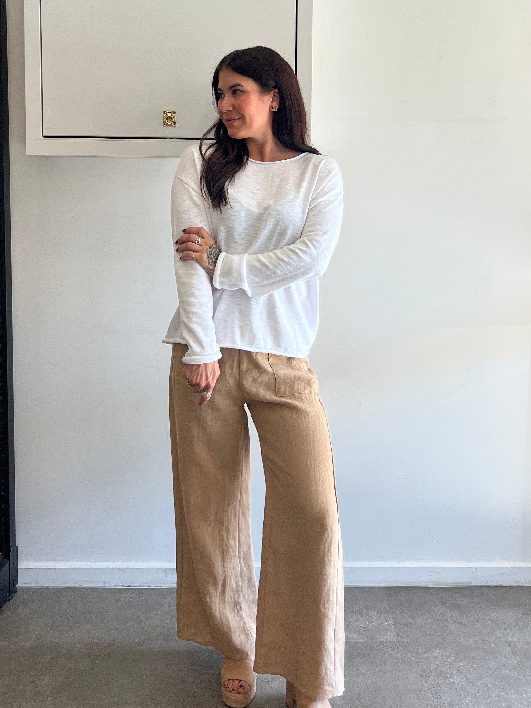 Woman wearing a white long-sleeve top and beige pants against a white wall.
