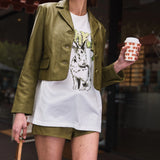 Woman in green jacket and white shirt with graphic design, holding a coffee cup.