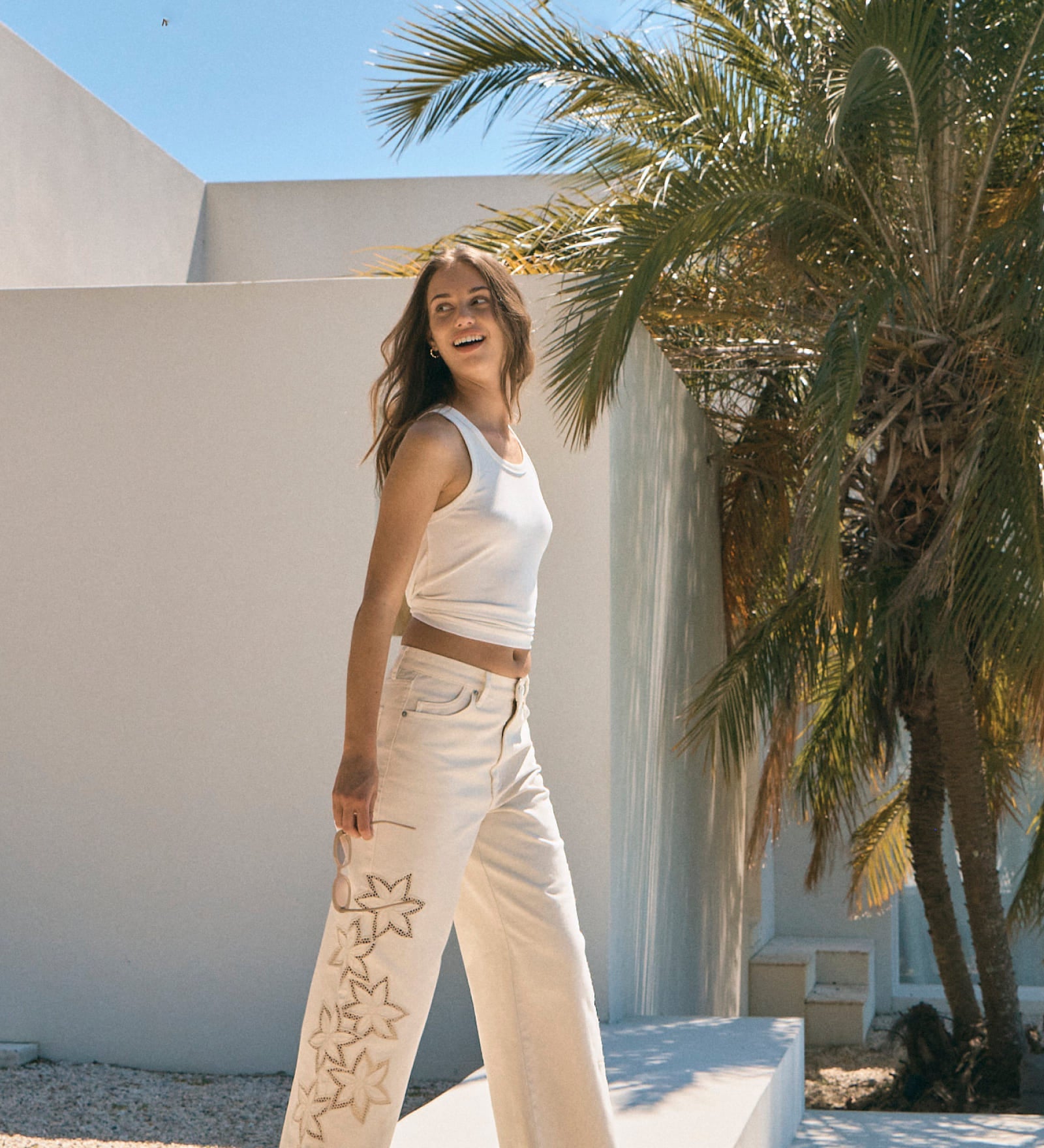 Woman in a white outfit standing in front of a palm tree and white wall.