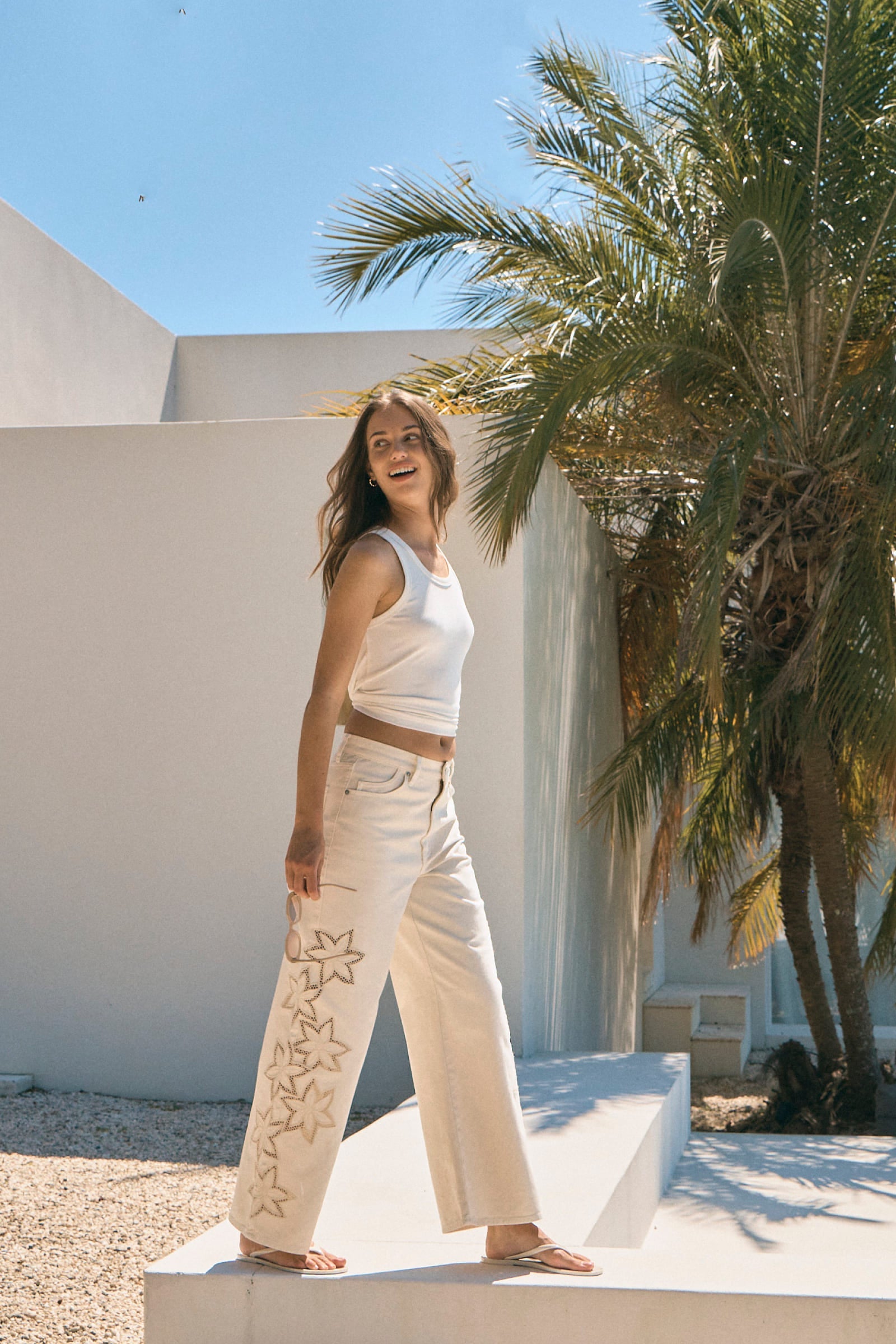 Woman in a white outfit standing in front of a palm tree and white wall.