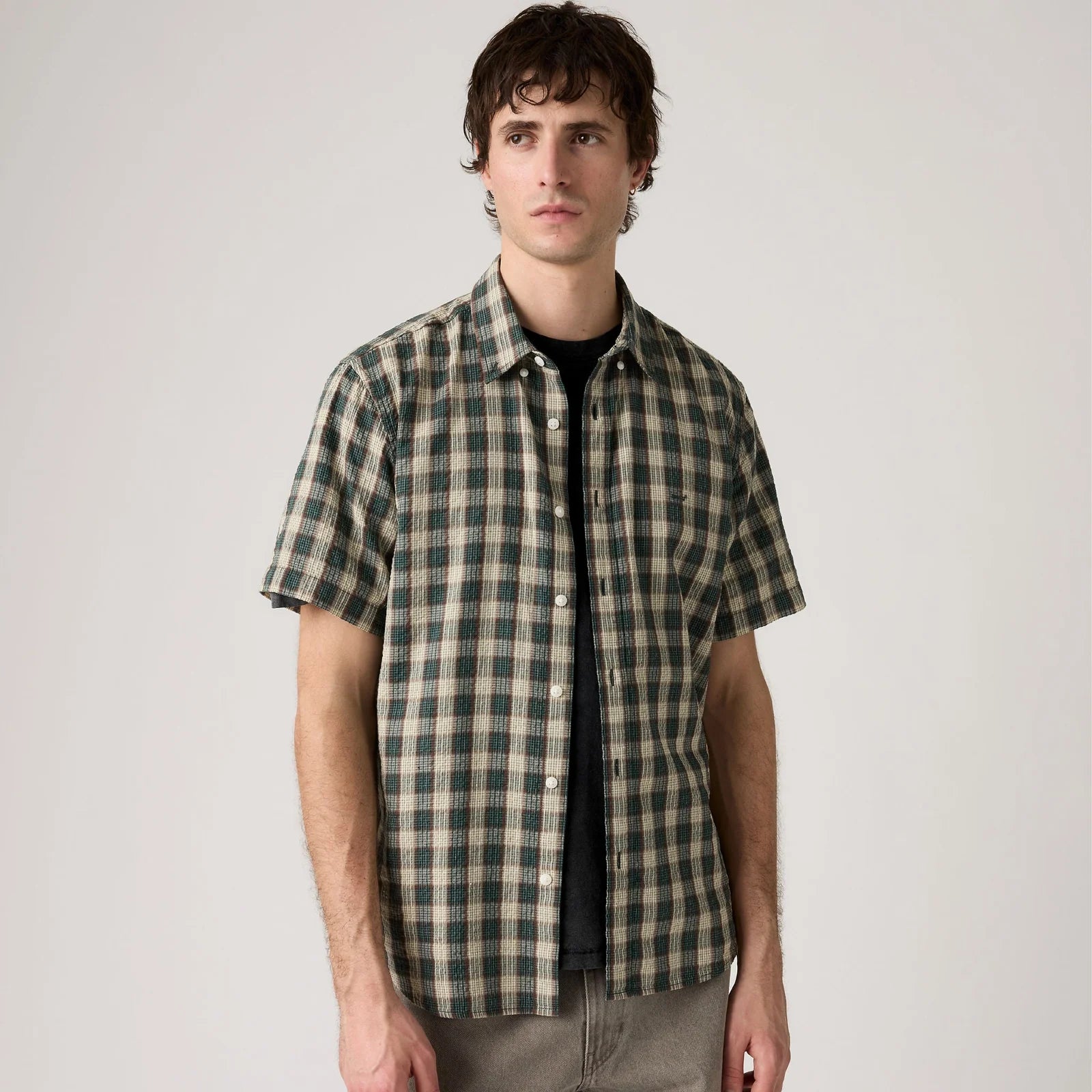 A man wearing a short-sleeve button-up plaid shirt in green and beige over a black t-shirt, standing against a plain light gray studio background.