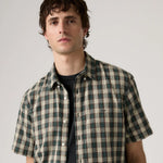A man wearing a short-sleeve button-up shirt with a green and beige plaid pattern over a black t-shirt, photographed against a neutral light gray background.
