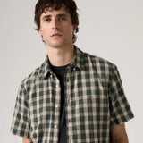 A man wearing a short-sleeve button-up shirt with a green and beige plaid pattern over a black t-shirt, photographed against a neutral light gray background.