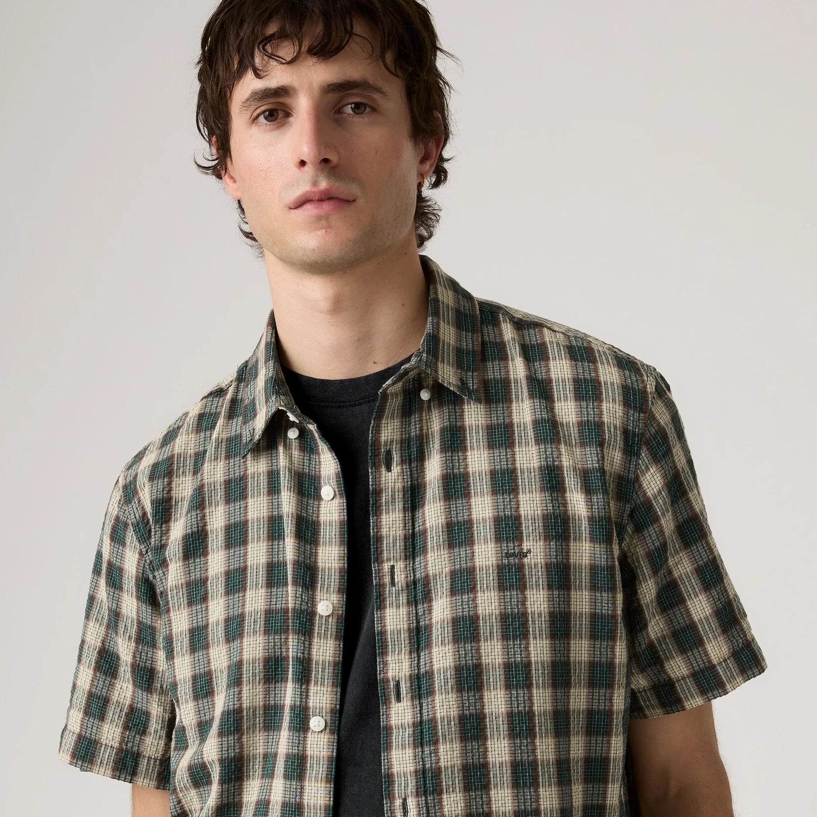 A man wearing a short-sleeve button-up shirt with a green and beige plaid pattern over a black t-shirt, photographed against a neutral light gray background.